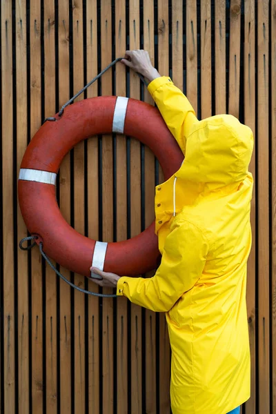 Man in a yellow raincoat hangs a red lifebuoy on a slatted wooden wall in the rain weather, back view. Outdoor.