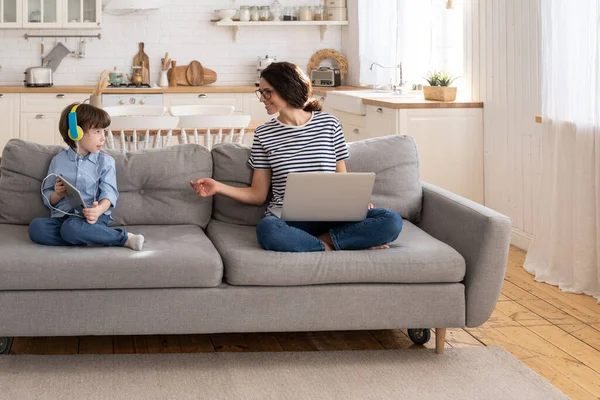 Mom freelancer remote work from home office on laptop sit on couch child playing on tablet. Lockdown