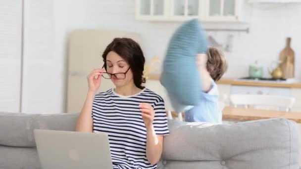 Erschöpfte Mutter sitzt zu Hause auf Sofa, arbeitet am Laptop, Kind lenkt ab, greift Mutter mit Kissen an — Stockvideo