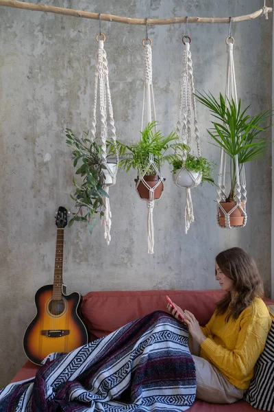Woman resting lying on sofa under cotton macrame plant hanger with houseplants using cellphone