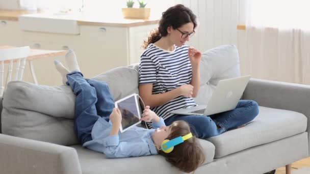 Мама фрілансер віддалена робота з домашнього офісу на ноутбуці сидить на дивані дитина грає на планшеті. Зафіксувати — стокове відео