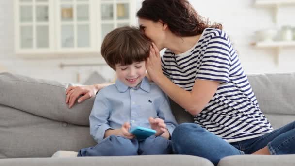 Moeder met kinderen op de bank genieten van grappige video op mobiele telefoon fluistert in oor, tijd doorbrengen thuis. — Stockvideo