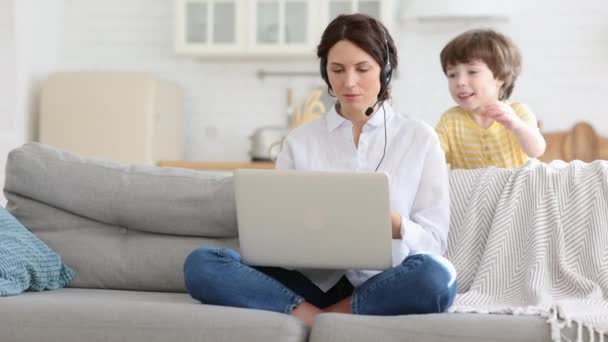 Exhausted mother sitting on couch at home, working on laptop, child distracted and asking attention — Stock Video