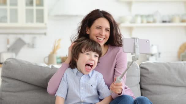 Matka a dítě drží telefon na selfie hůl pro video hovor ukazuje jazyk sedí na gauči doma — Stock video