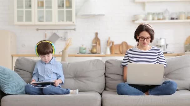 Mamma frilansare fjärrarbete från hemmakontoret på laptop sitta på soffa barn leker på surfplatta. Nedstängning — Stockvideo