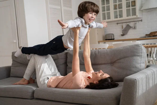 Happy loving mother with cute son imitate plane lying on couch in living room in morning on weekend — Stock Photo, Image