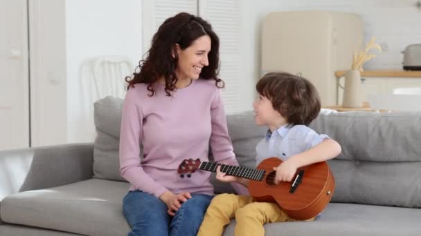 Söt förskolekille hålla ukulele, skratta sitter tillsammans med mamma i vardagsrummet. Fritid för familjer — Stockvideo