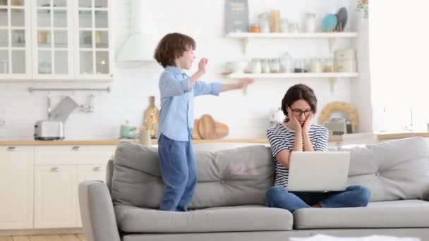 Trött mamma sitta på soffan, arbeta på laptop hemma, hyperaktiv liten unge hoppar uppmärksamhet. — Stockvideo