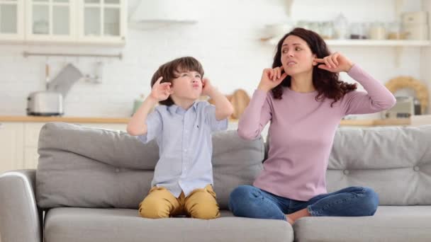 Mãe franzina e filho sentam-se no sofá com os olhos fechados e cobrem os ouvidos de música barulhenta ou sons de luta — Vídeo de Stock