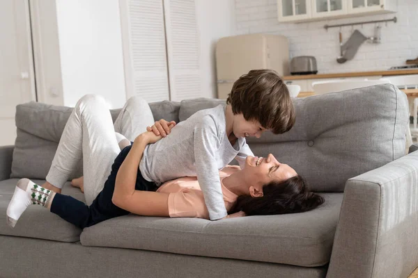 Überglückliche verspielte Mutter und Sohn umarmen sich, lachen am Wochenendmorgen zu Hause auf dem Sofa im Wohnzimmer liegend — Stockfoto