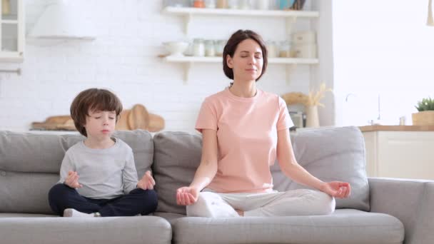 Mama z uroczym synkiem ćwiczy jogę siedzieć razem na kanapie w domu ucząc dziecko medytować — Wideo stockowe
