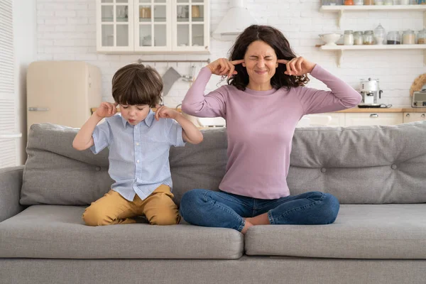 Fruncir el ceño mamá y el hijo se sientan en el sofá con los ojos cerrados y cubren los oídos de la música ruidosa o sonidos de lucha —  Fotos de Stock