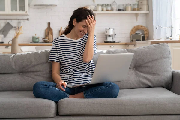 Vrolijke jonge vrouw lachen terwijl kijken grappige video op laptop thuis zitten met gekruiste benen op de bank — Stockfoto