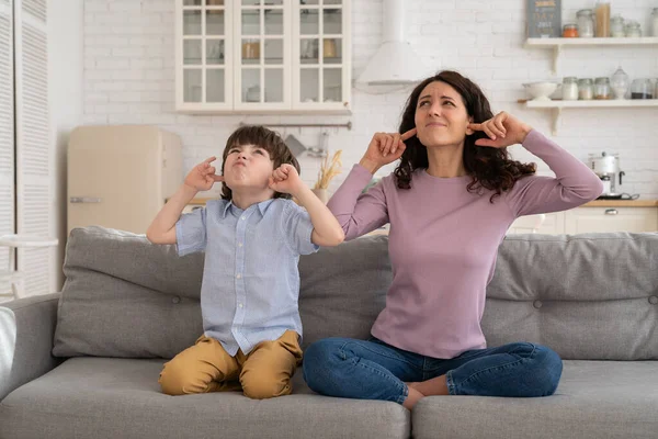 Frowning mom and son sit on couch with closed eyes and cover ears from noisy music or fight sounds