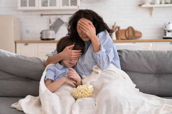 Mom and kid son with bowl of popcorn watching scary movie closing their eyes sitting on sofa at home