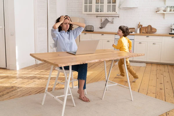 Freelance werknemer vrouw moe van luidruchtige zoon werk op laptop computer in de keuken met kleine zoon draait — Stockfoto