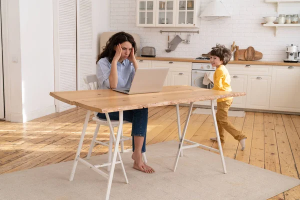 Gefrustreerde moeder werken op afstand op laptop in huis kantoor zitten aan keukentafel en luidruchtige zoon lopen — Stockfoto