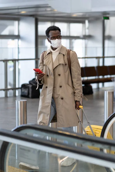 Schwarzer Reisender mit Koffer geht zur Rolltreppe im Flughafenterminal, trägt Gesichtsmaske. Neue Normalität. — Stockfoto