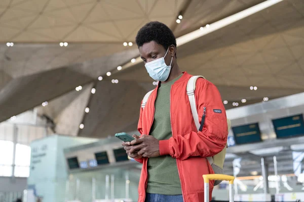 Young black male traveler in airport wait for flight use smartphone wear mask in covid-19 epidemic