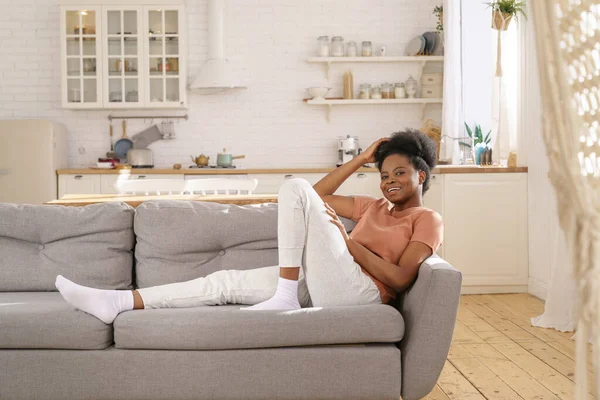 Glückliche junge schwarze Frau liegt entspannt auf dem Sofa. Afroamerikanerin in Kamera mit zahmem Lächeln — Stockfoto