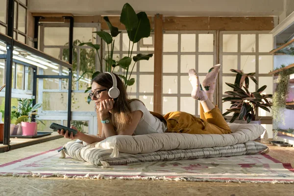 Chica jardinero relajarse después del trabajo yacía en el suelo, mensaje en el teléfono inteligente y escuchar música. Inicio relajación — Foto de Stock