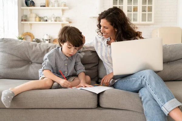 Home school: smiling mom help little son to write in notebook during rest of remote work on laptop