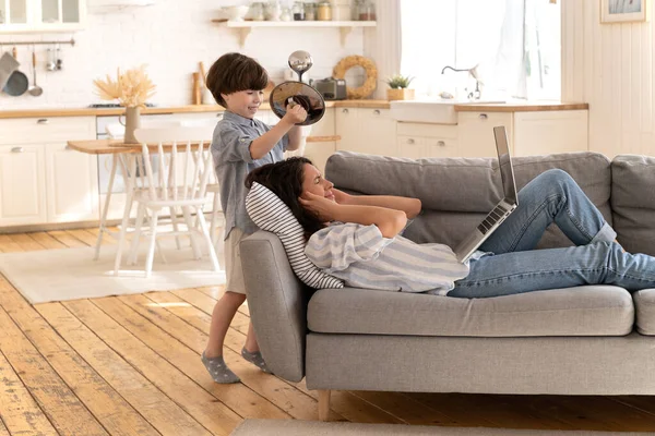 Stressed mom cover ear from noisy son bang dishes. Tired of kid woman lying with closed eyes on sofa — Stock Photo, Image