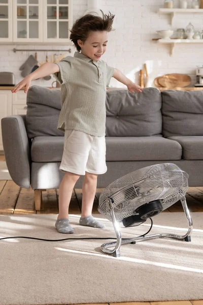Joyful kleine jongen alleen in de woonkamer genieten van frisse wind uit retro ventilator of airconditioner — Stockfoto