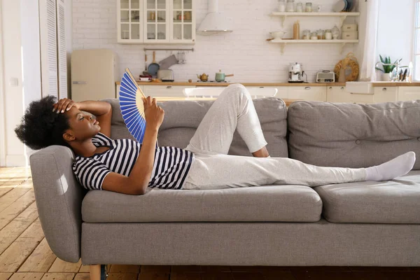 Femme noire souffrant de coup de chaleur plat sans climatiseur hochant ventilateur couché sur le canapé à la maison — Photo