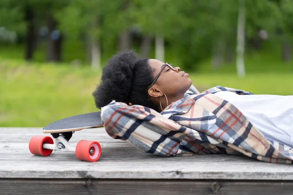 Mujer joven africana relajada acostada en longboard con los ojos cerrados soñando despierto al aire libre en el monopatín — Foto de Stock