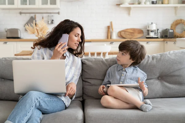 Femme d'affaires occupée mère travail de la maison parler à l'enfant avec ordinateur portable sur les genoux appelant sur le téléphone sur le canapé — Photo