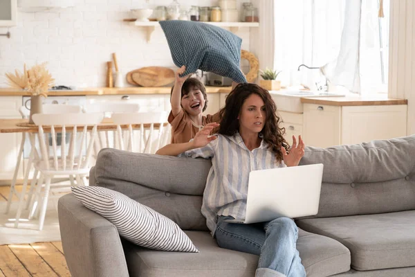 Boos jong mam proberen te concentreren op het werk in laptop als kleine ongehoorzame kind sloeg haar met kussen — Stockfoto