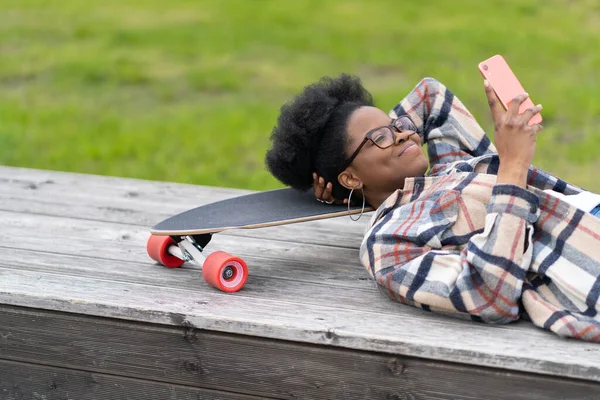 웃는 스케이트타는 여자는 긴 칠판에 누운 채 휴식을 취하거나 즐거운 미소를 띠고 스마트폰으로 메시지를 읽는다 — 스톡 사진
