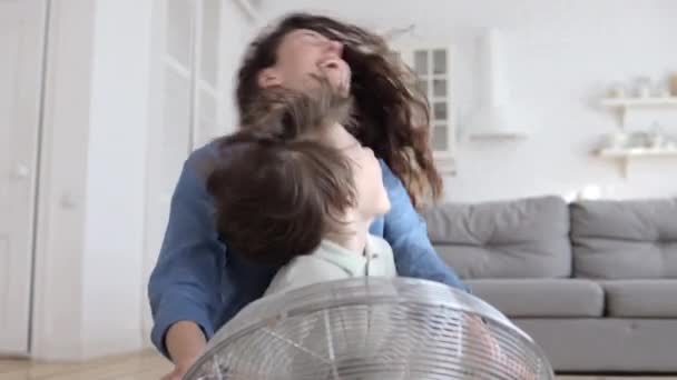Kleiner Sohn und freudige Mama haben Spaß mit großem Ventilator, der im Sommer im Wohnzimmer auf dem Boden sitzt — Stockvideo