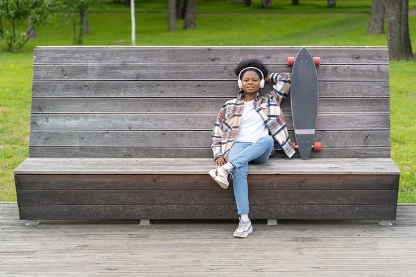 Χαλαρός αφρικανή γυναίκα skateboarder chill ακούσετε μουσική με ακουστικά και longboard στο αστικό χώρο — Φωτογραφία Αρχείου