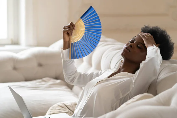 Schwarze Geschäftsfrau, die an Hitzschlag leidet, sitzt mit winkendem Ventilator im heimischen Wohnzimmer — Stockfoto
