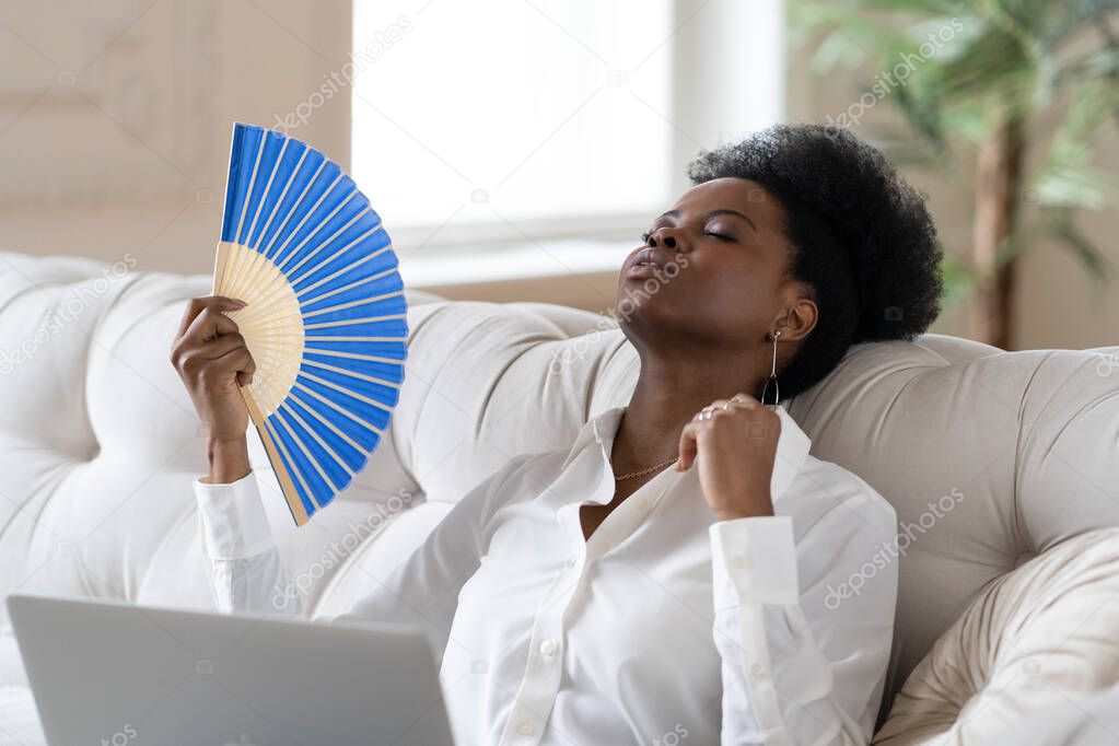 Black business woman suffering from heat stroke sitting in living room at home using waving fan