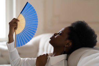 Black business woman suffering from heat stroke sitting in living room at home using waving fan clipart