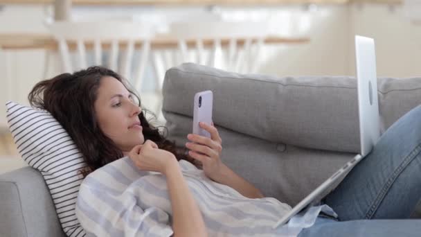 Trabalho freelancer feminino jovem deitado no sofá com computador portátil nos joelhos e smartphone nas mãos — Vídeo de Stock