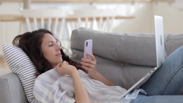 Jonge vrouwelijke freelancer werk liggend op de bank met laptop computer op knieën en smartphone in handen — Stockvideo