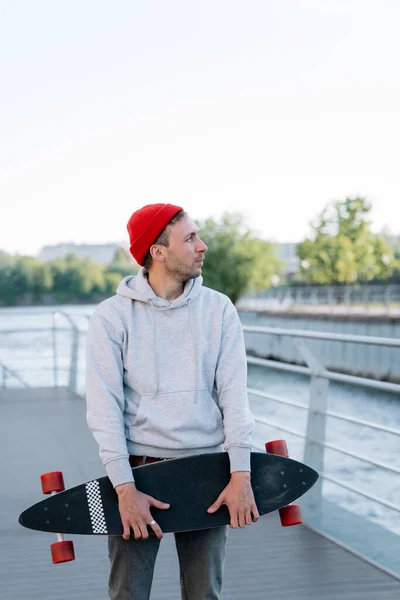 Homem jovem hipster relaxado skatista segurar longboard na ponte ao pôr do sol. Conceito de estilo de vida urbano — Fotografia de Stock