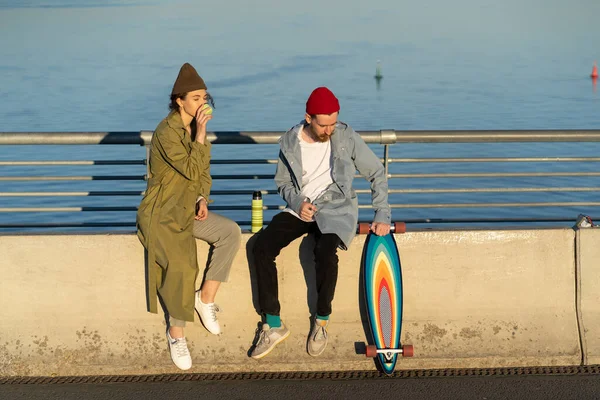 Casual hipsters τύπος και κορίτσι σε ημερομηνία: μαζί πίνουν τσάι κάθεται στη γέφυρα πάνω από το ποτάμι στον ήλιο ηλιοβασιλέματος — Φωτογραφία Αρχείου