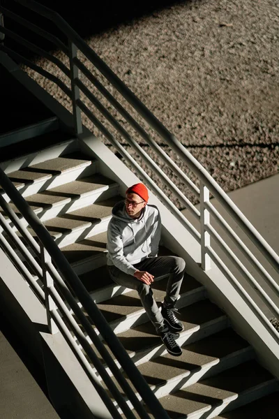 Самотній молодий чоловік в окулярах сидить наодинці на бетонних сходах у міському будівництві або міському просторі, зверху — стокове фото