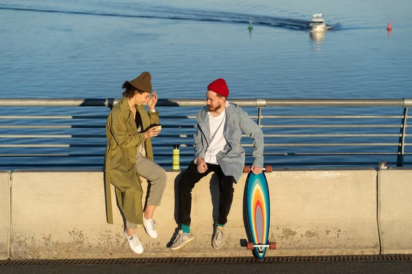 Пара закоханих хіпстерів сидить на бетонній тротуарі річкового мосту розмовляють і сміються пити гарячий чай — стокове фото