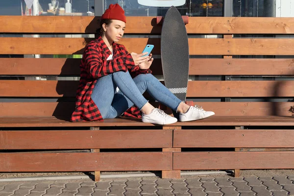 Módní ležérní hipster dívka s chytrým telefonem sedí na lavičce venku s longboard stojí v blízkosti — Stock fotografie