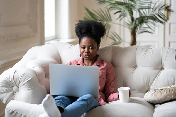 Serious african woman work on laptop sit on sofa with coffee or tea relaxing after remote occupation — Photo