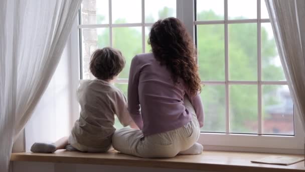 Mãe e filho sentam-se no peitoril da janela olhar na janela abraçando abraçar juntos em casa na quarentena covid-19 — Vídeo de Stock