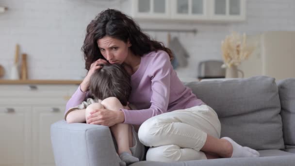 疼爱的母亲抱着心烦意乱的小儿子坐在家里的沙发上，母亲抚慰着被冒犯的伤心学龄前男孩 — 图库视频影像