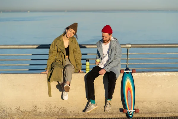 Trendy couple enjoy sunset sun with tea and longboard on concrete bridge dressed in street clothes