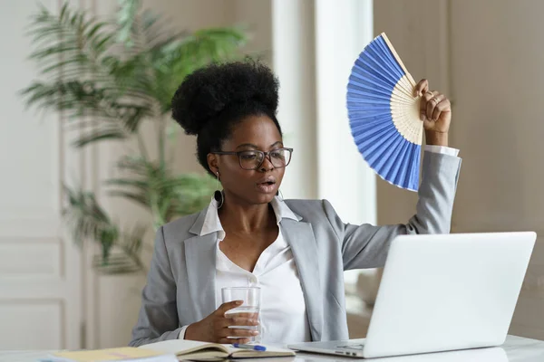 Überhitzte afrikanische Geschäftsfrau winkt Ventilator trinken Wasser sitzt am Schreibtisch mit Laptop oder zu Hause — Stockfoto